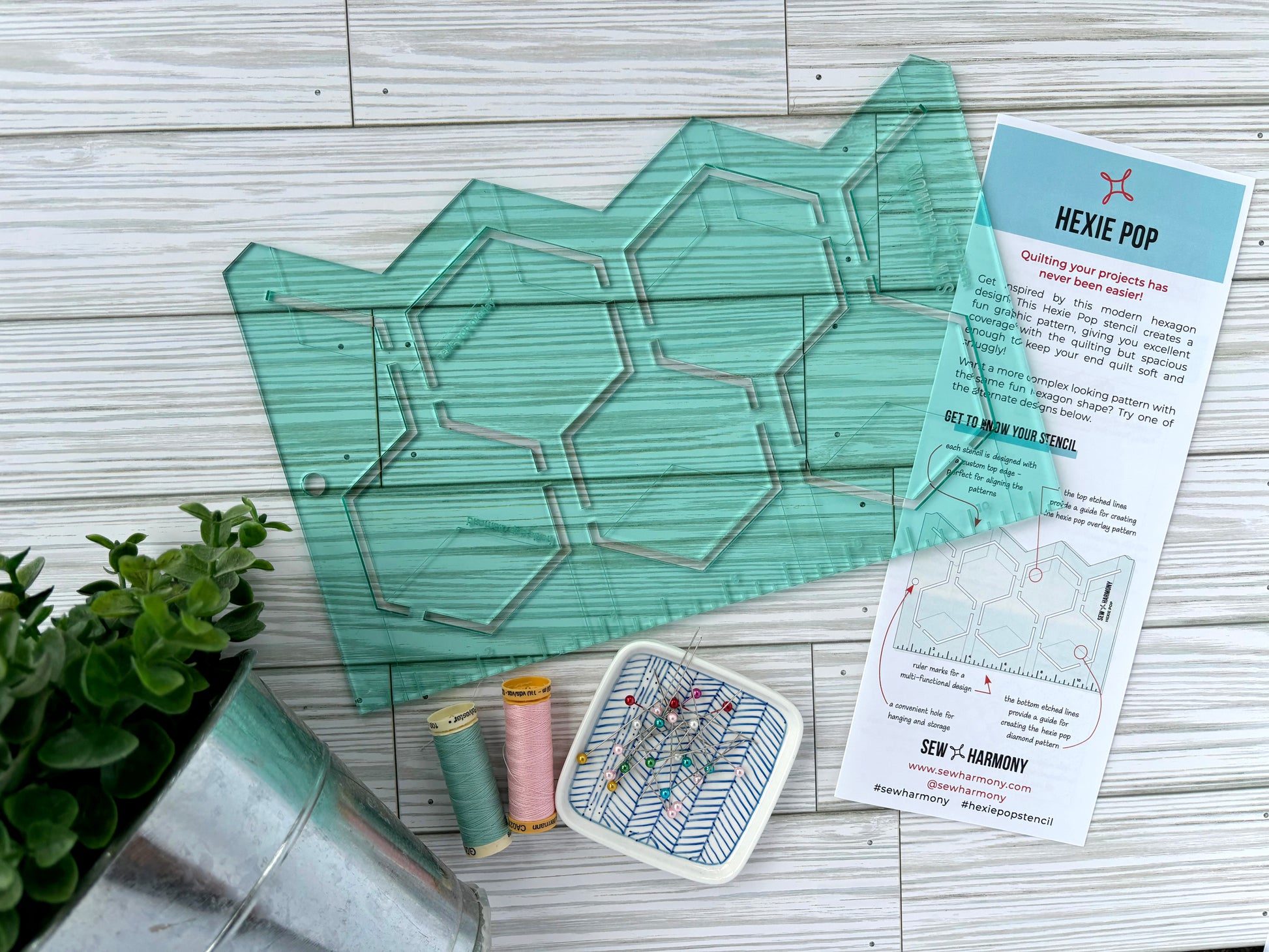 A light green transparent acrylic quilting stencil with a hexagon pop shaped design. Pictured with the stencil is a small plant, two spools of thread, a dish of pins, and an information pamphlet. This is all displayed on a white-washed woodgrain surface.