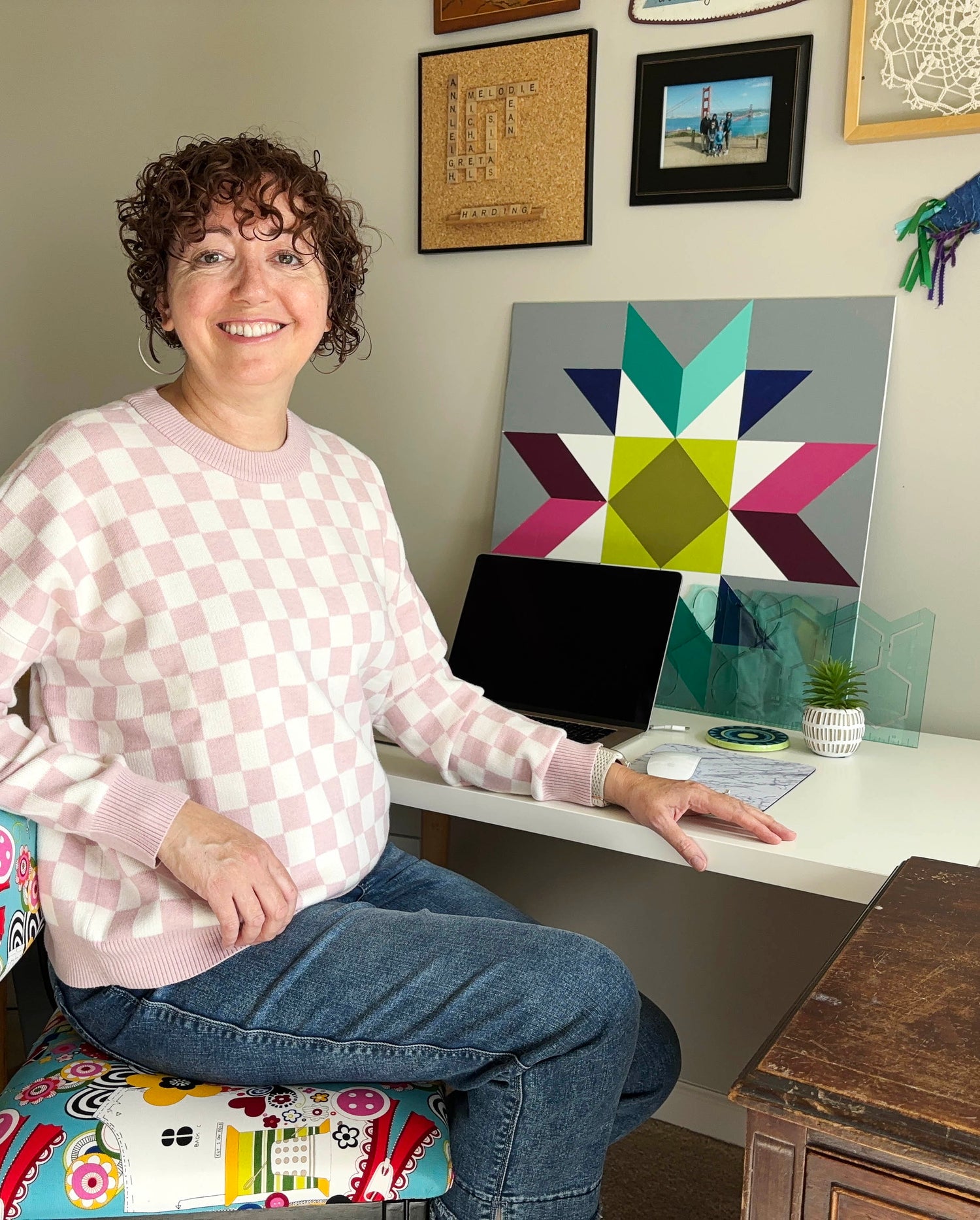 A photo of a woman sitting at a desk near a computer and a barn quilt.