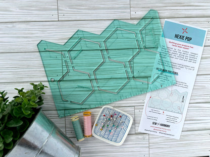 A light green transparent acrylic quilting stencil with a hexagon pop shaped design. Pictured with the stencil is a small plant, two spools of thread, a dish of pins, and an information pamphlet. This is all displayed on a white-washed woodgrain surface. 