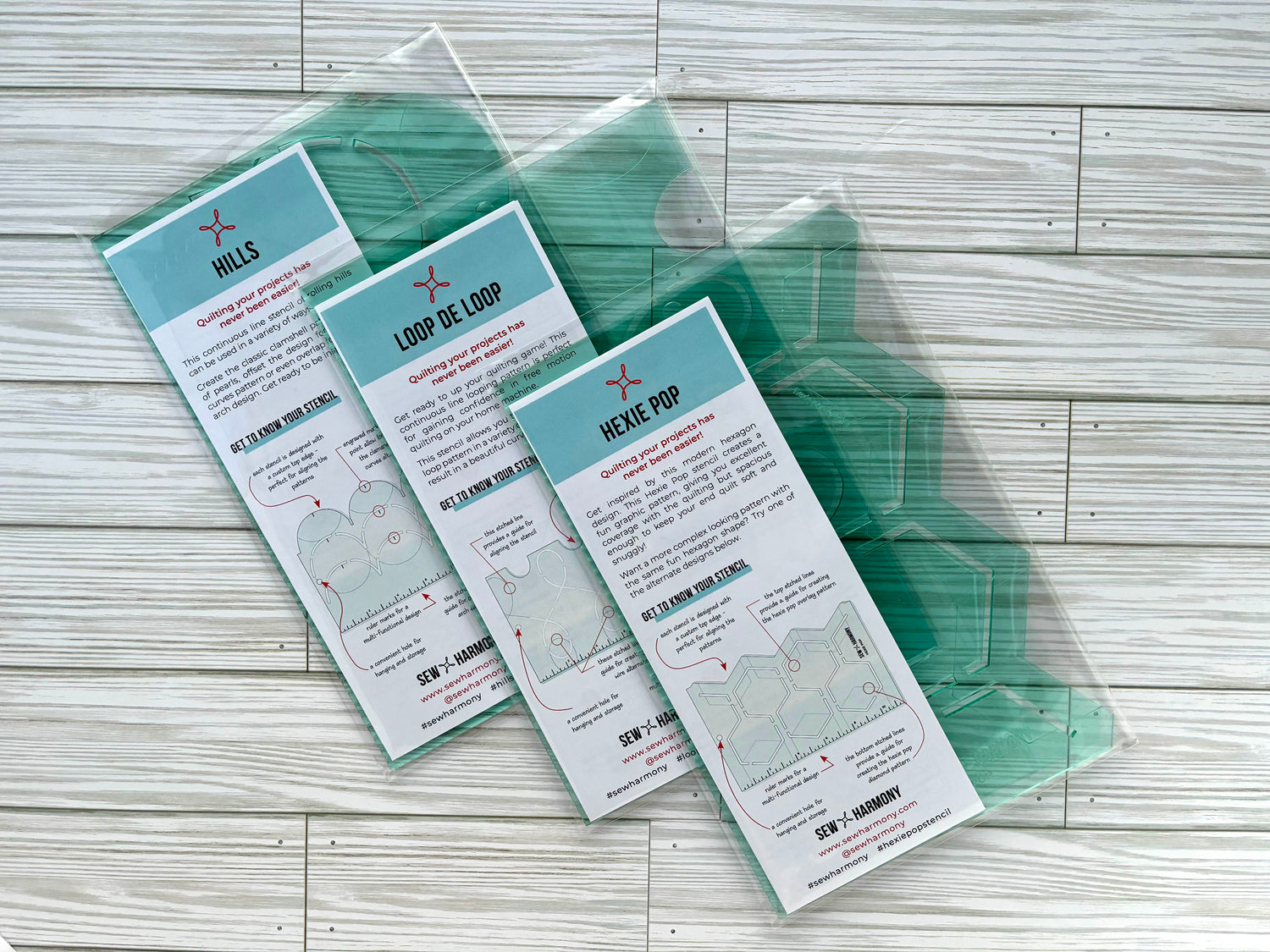 SEW * HARMONY's three signature quilting stencils packaged with their information pamphlets and displayed on a white-wash woodgrain surface. The three quilting stencils are the Hexie Pop, Loop De Loop, and Hills acrylic stencils made from 3mm transparent light green material.