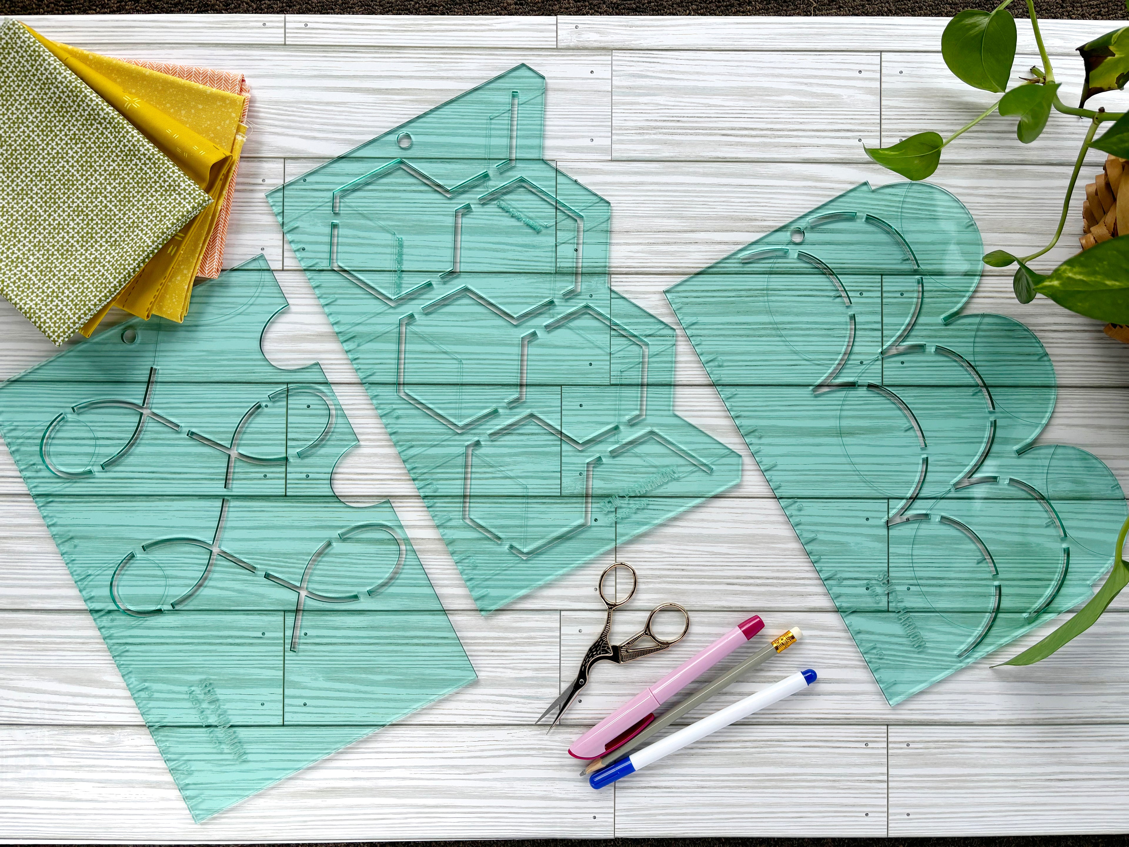 An image of SEW * HARMONY's three signature quilting stencils. The Loop De Loop, Hexie Pop, and Hills stencils. These 3mm acrylic stencils are made from transparent light green material and are shown on a whitewash wood surface surrounded by a stack of fabric, plant, three marking tools and a pair of embroidery scissors. 