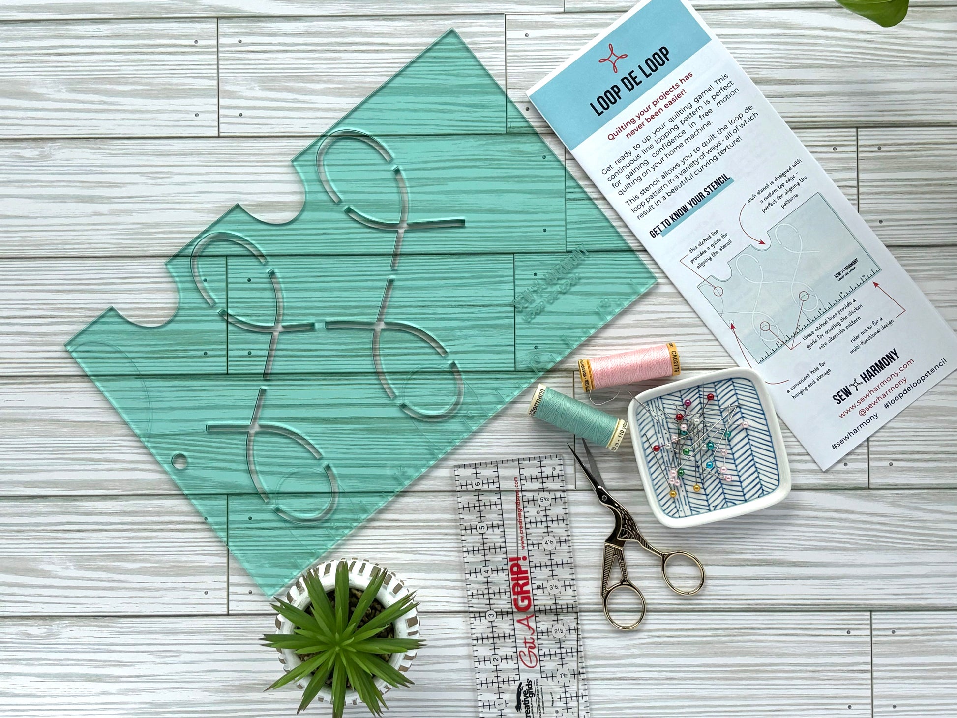 A transparent light green acrylic stencil featuring a looping pattern accompanied by an instruction pamphlet, ruler, scissors, thread, bowl of pins and small plant.