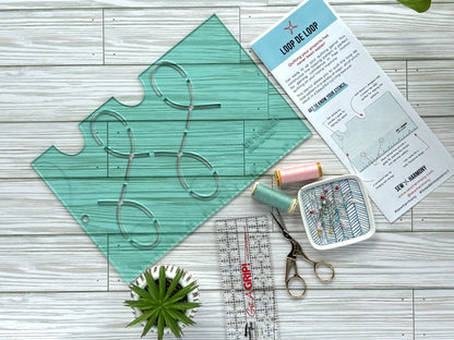 A transparent light green acrylic stencil featuring a looping pattern accompanied by an instruction pamphlet, ruler, scissors, thread, bowl of pins and small plant.